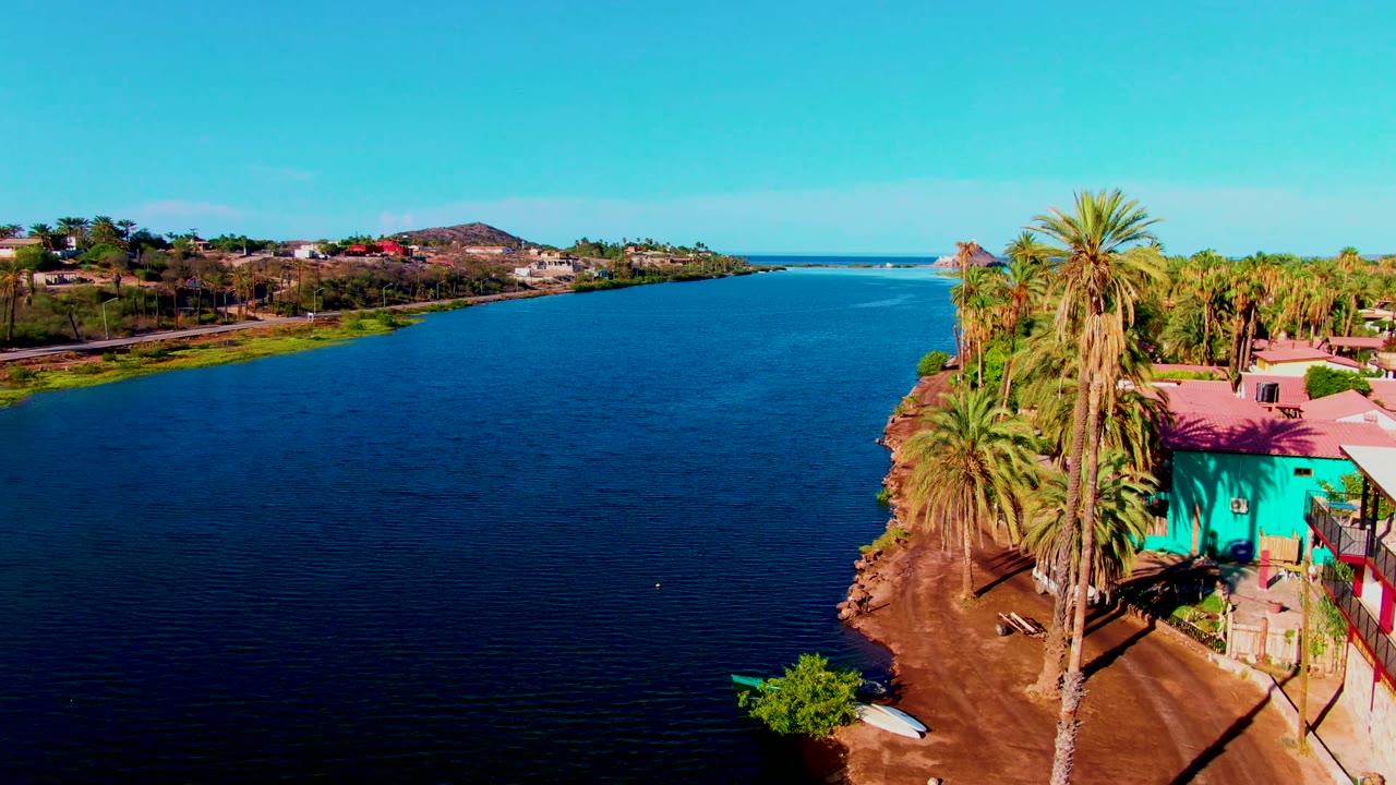 Aerial view of the Mulege riverfront with the turquoise building and palm trees more prominent at frame right, rippled water across the foreground, homes on the far bank, and no visible people.