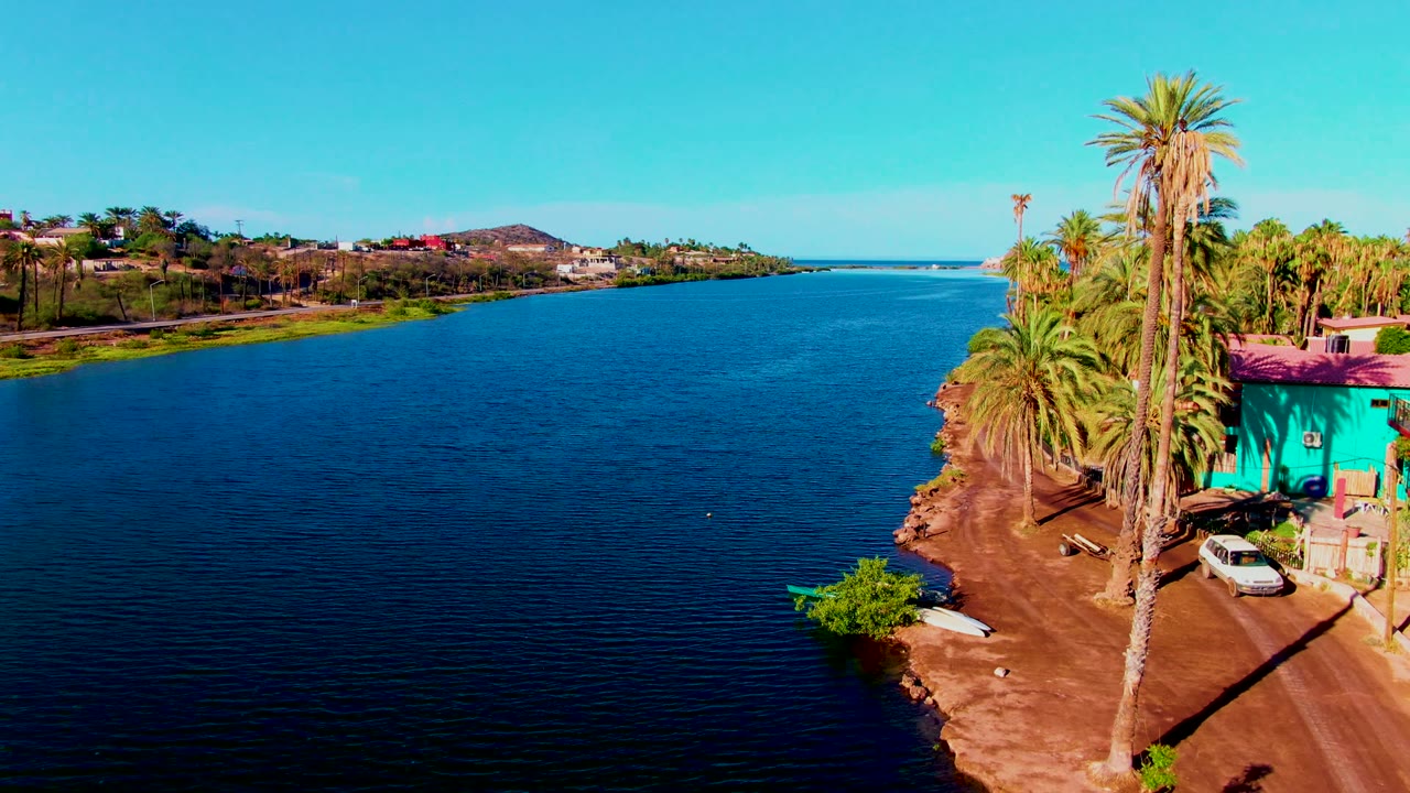 Aerial frame from Mulege, Baja California Sur showing the blue canal on the left, palm trees and a turquoise shoreline building on the right, a white car near the dirt road, and no visible people.