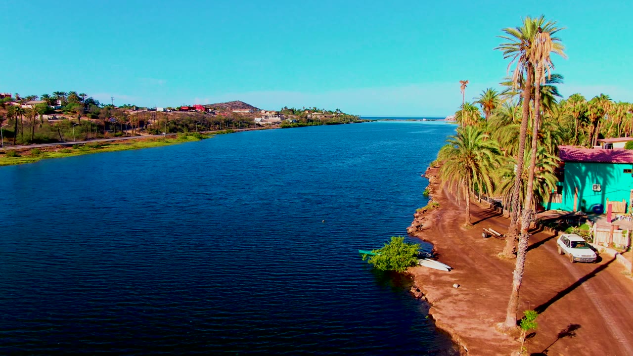 Wide aerial canal view with palm-lined dirt shore, turquoise building, and a white car near the right edge.