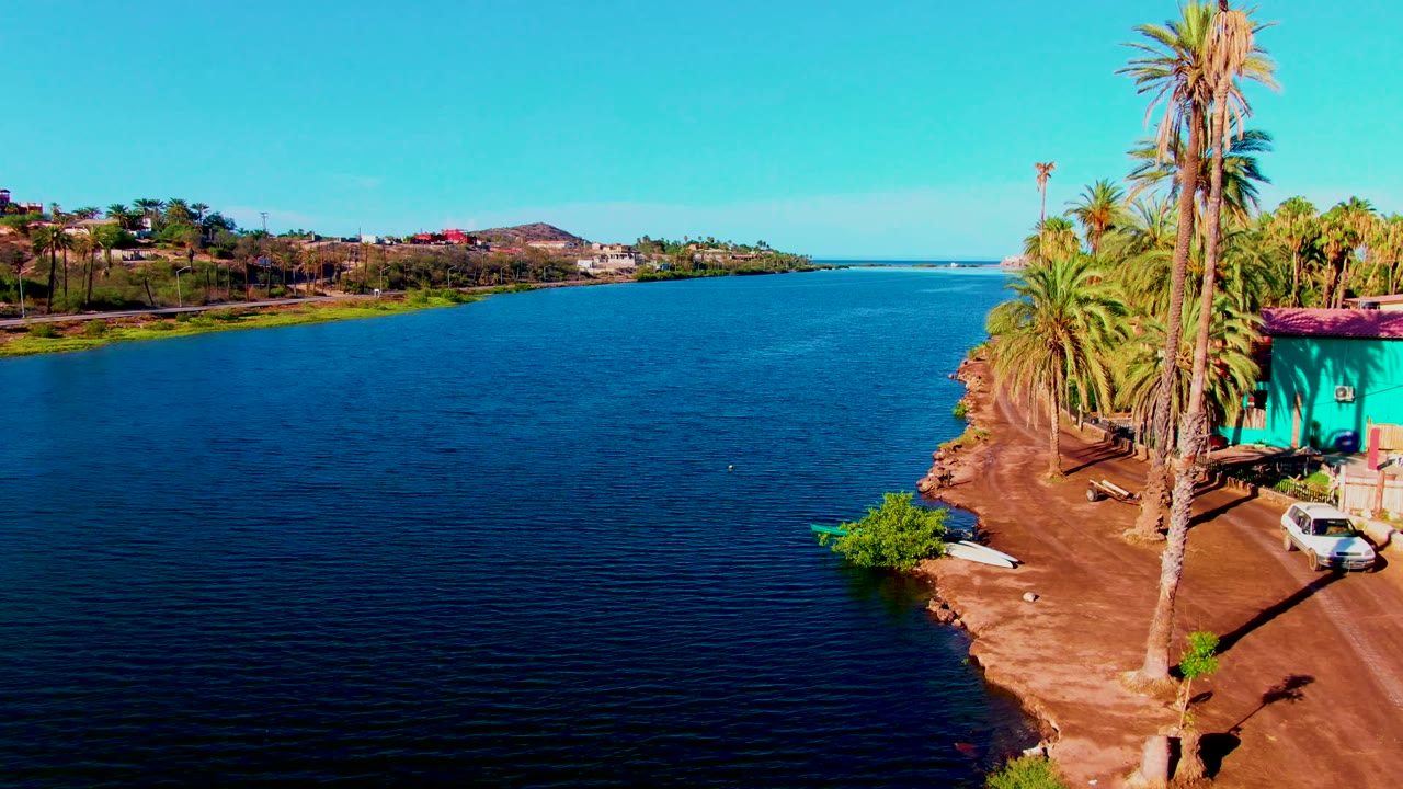 The 0:02 frame shows rippled blue canal water in Mulege, Baja California Sur, with the dirt shoreline, tall palms, turquoise building, white car, and no visible people along the right edge.