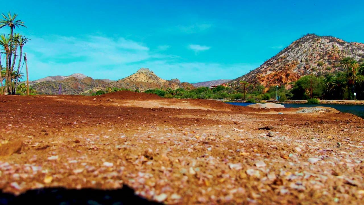A low Mulege lakeshore frame shows red dirt and scattered stones in front, palm clusters and a narrow lake edge to the side, rocky mountains beyond, and no visible people.