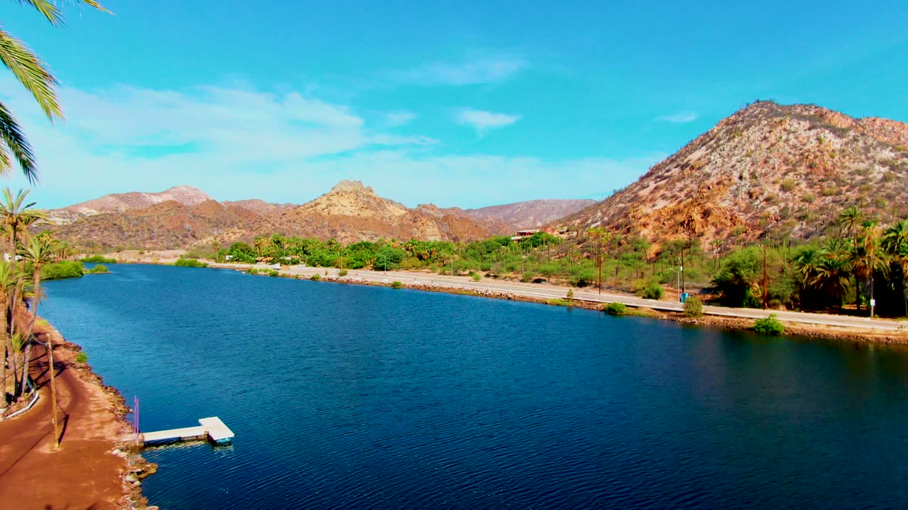 Opening aerial frame over blue water in Mulege, Baja California Sur, with a small dock at lower left, palms along the shore road, rocky mountains beyond, and no visible people.