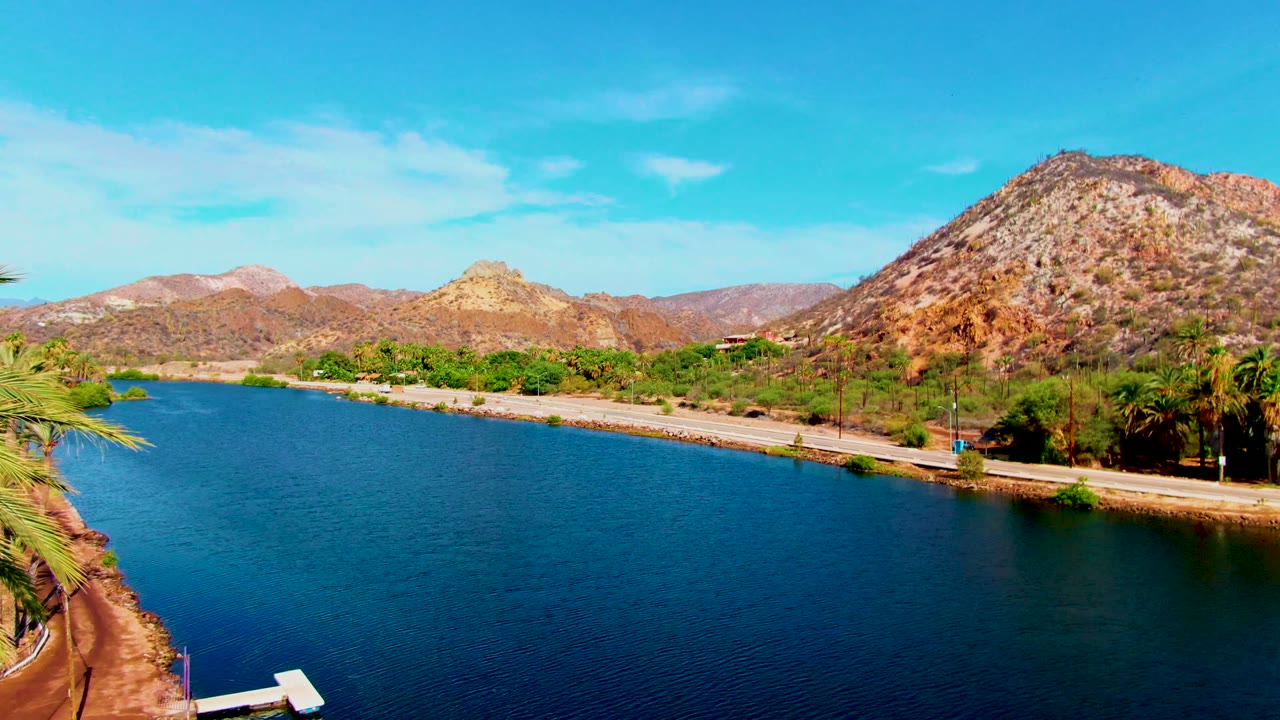 Blue water beside a shore road with palms and arid mountains behind it.