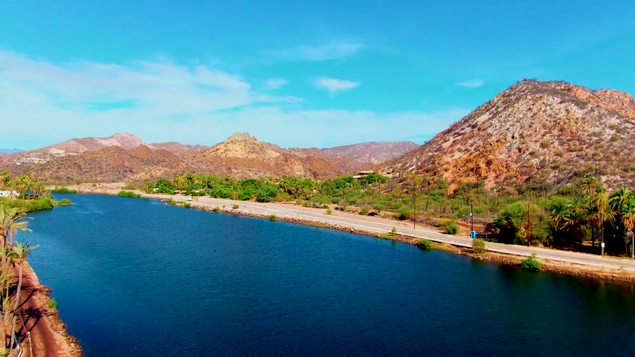 Mid-pan aerial view in Mulege, Baja California Sur with broad blue water in the foreground, a pale road along the shore, dense palms to the right, rocky hills, and no visible people.