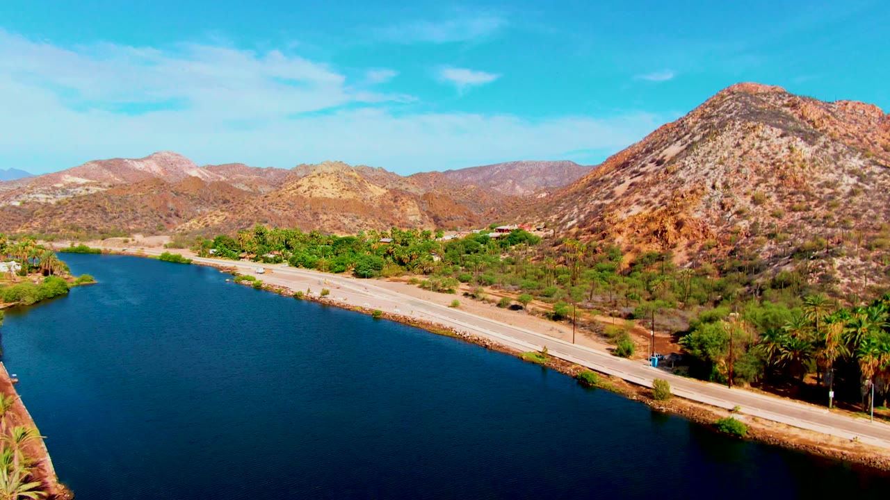 Wide aerial view in Mulege, Baja California Sur showing dark blue canal water beside a pale road, green roadside trees, rocky hills, bright sky, and no visible people.