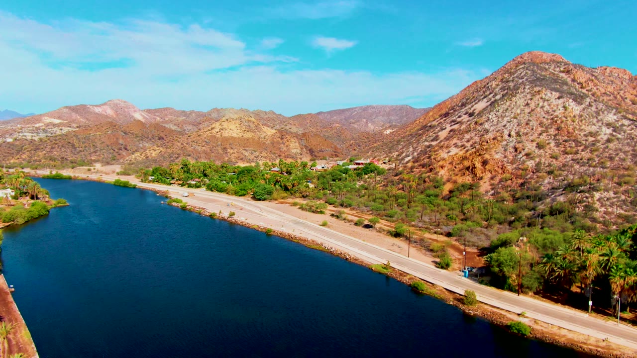 Mulege aerial view with deep blue water in the lower frame, an empty-looking road along the canal, green palms at the bank, reddish rocky hills to the right, and no visible people.