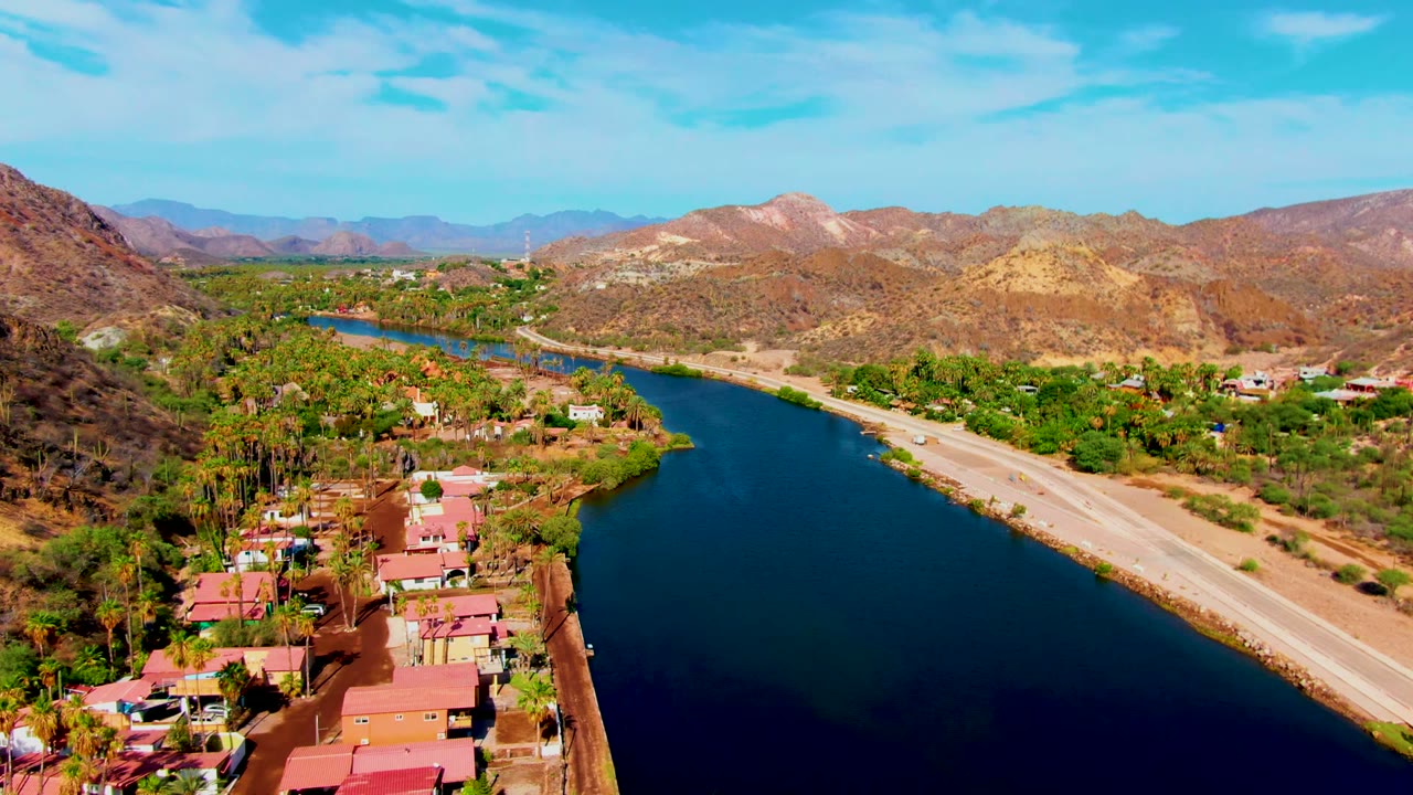 High aerial frame over Mulege, Baja California Sur showing blue canal water, a parallel road, red-roofed buildings, palms, dry mountains, clear sky, and no visible people.