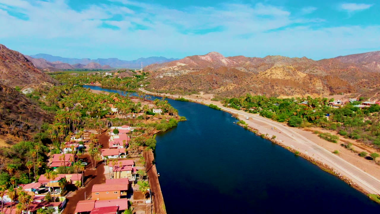 Aerial canal scene with red roofs, road, palms, greenery, and brown hills.