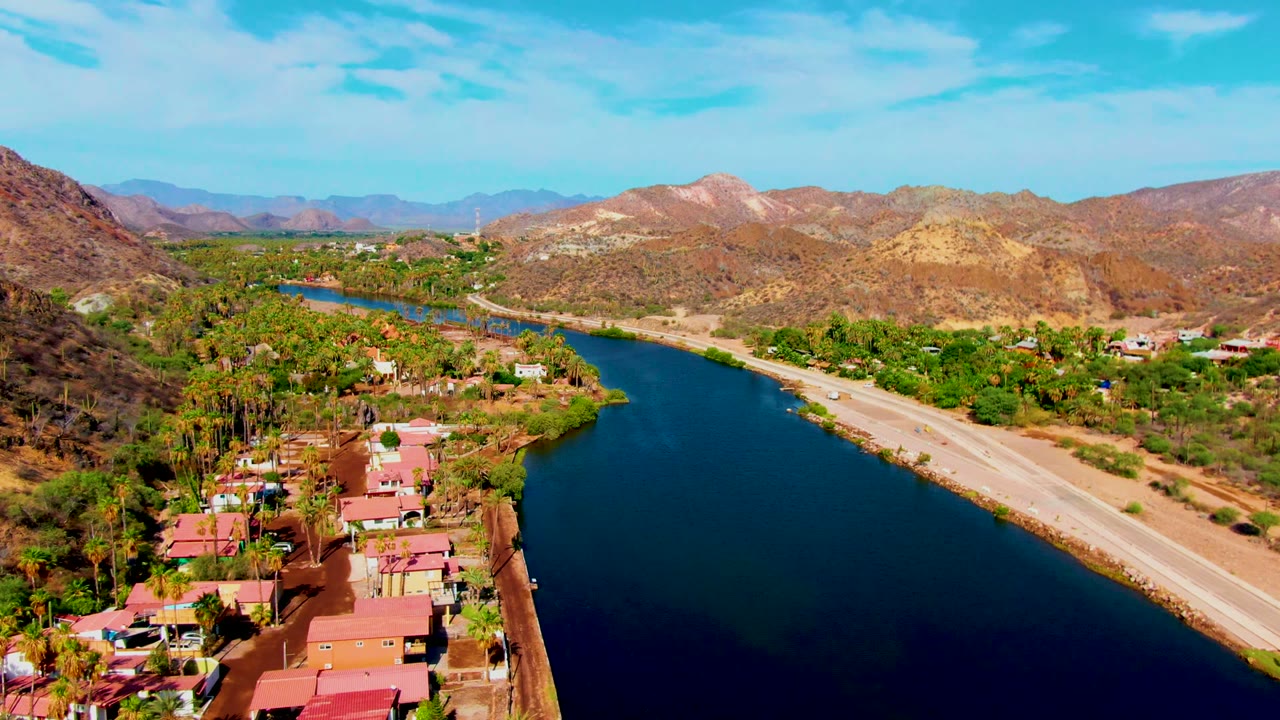 Aerial view over Mulege with deep blue canal water beside the roadway, compact buildings and trees near the bank, layered desert mountains, and no visible people.