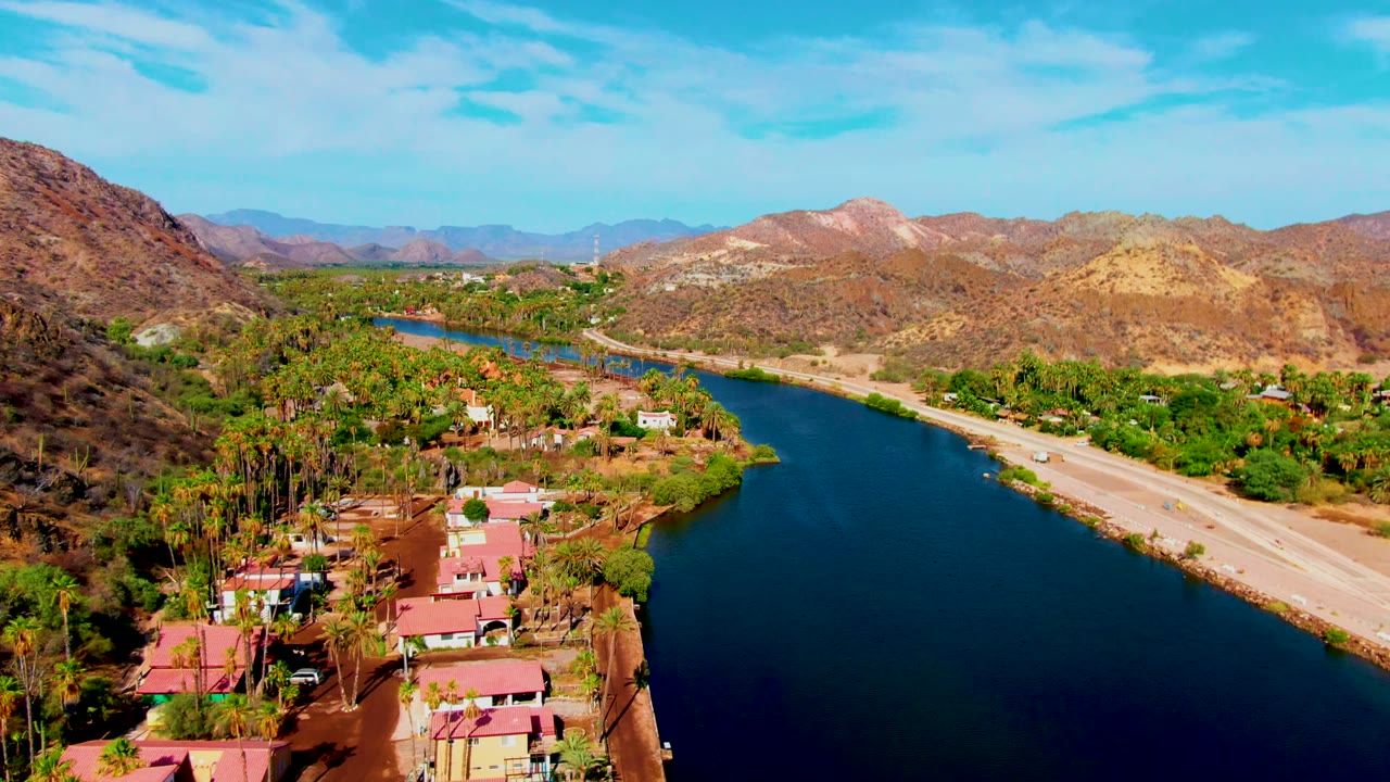 Aerial view over Mulege, Baja California Sur, showing a blue river beside a pale road, red-roofed homes, palms, dry hills, and no visible people.