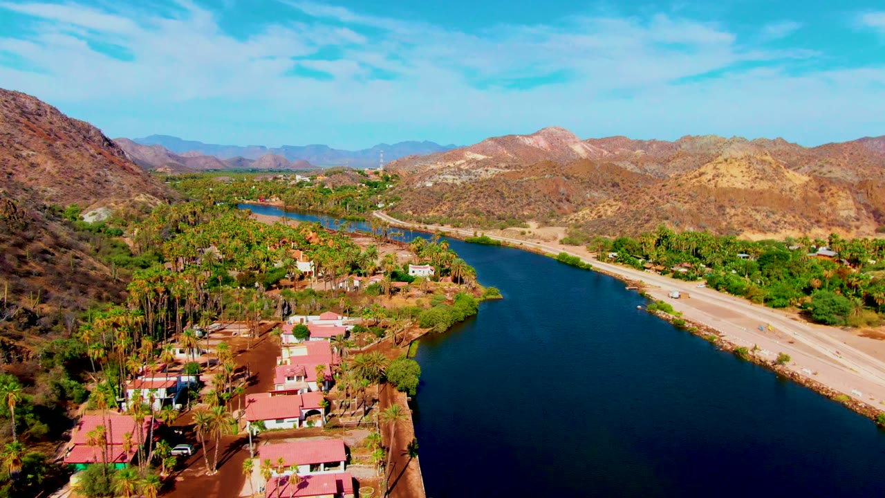 Wide aerial view of water, roadside traffic, palms, houses, and mountains.