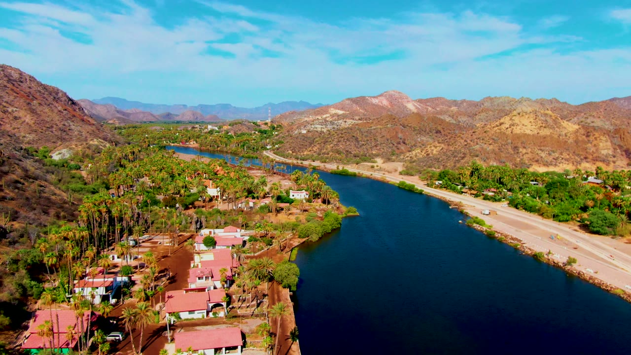 Aerial view in Mulege showing the dark blue waterway, a light road with small vehicles, dense green palms, red roofs, and layered arid mountains under clear sky, with no visible people.