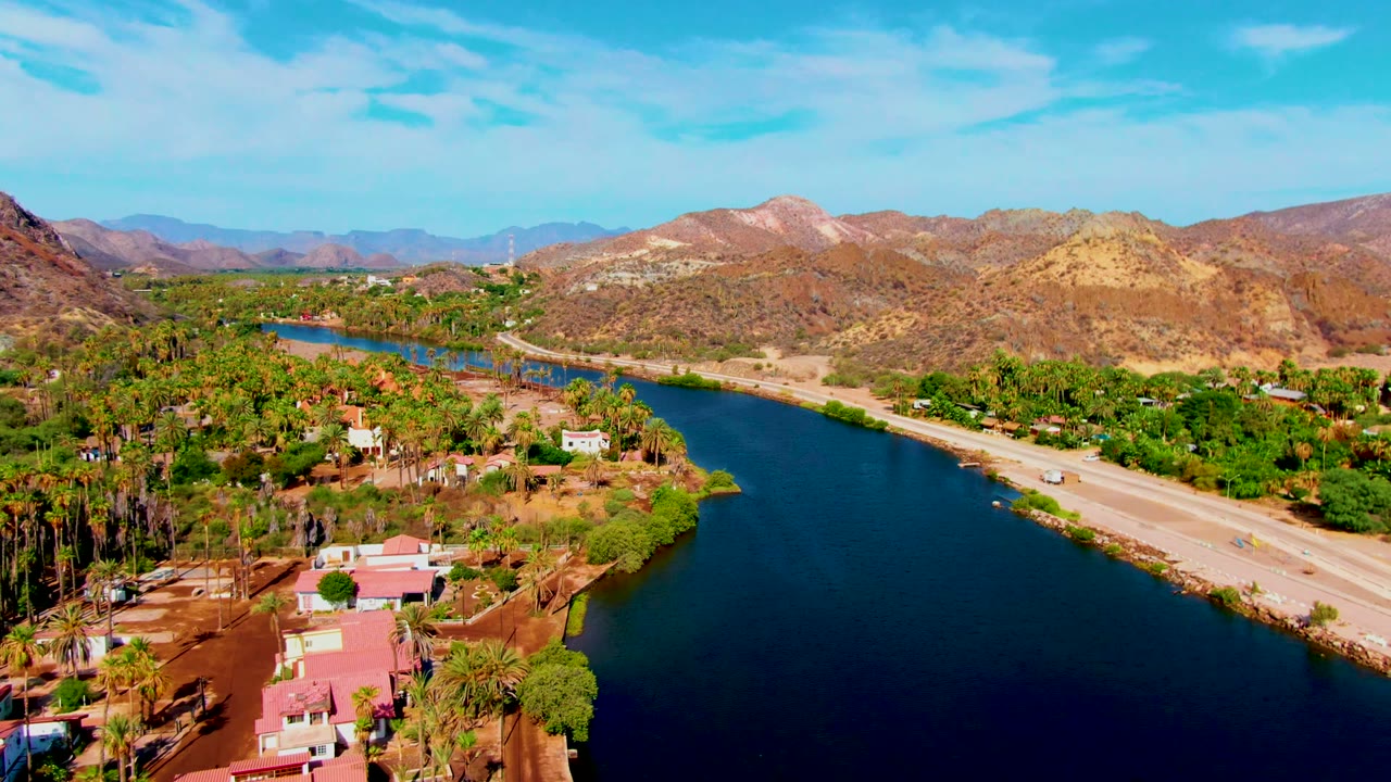 Opening aerial frame over Mulege, Baja California Sur, with blue river water in the foreground, a road along the bank, palms, low buildings, dry hills, and no visible people.