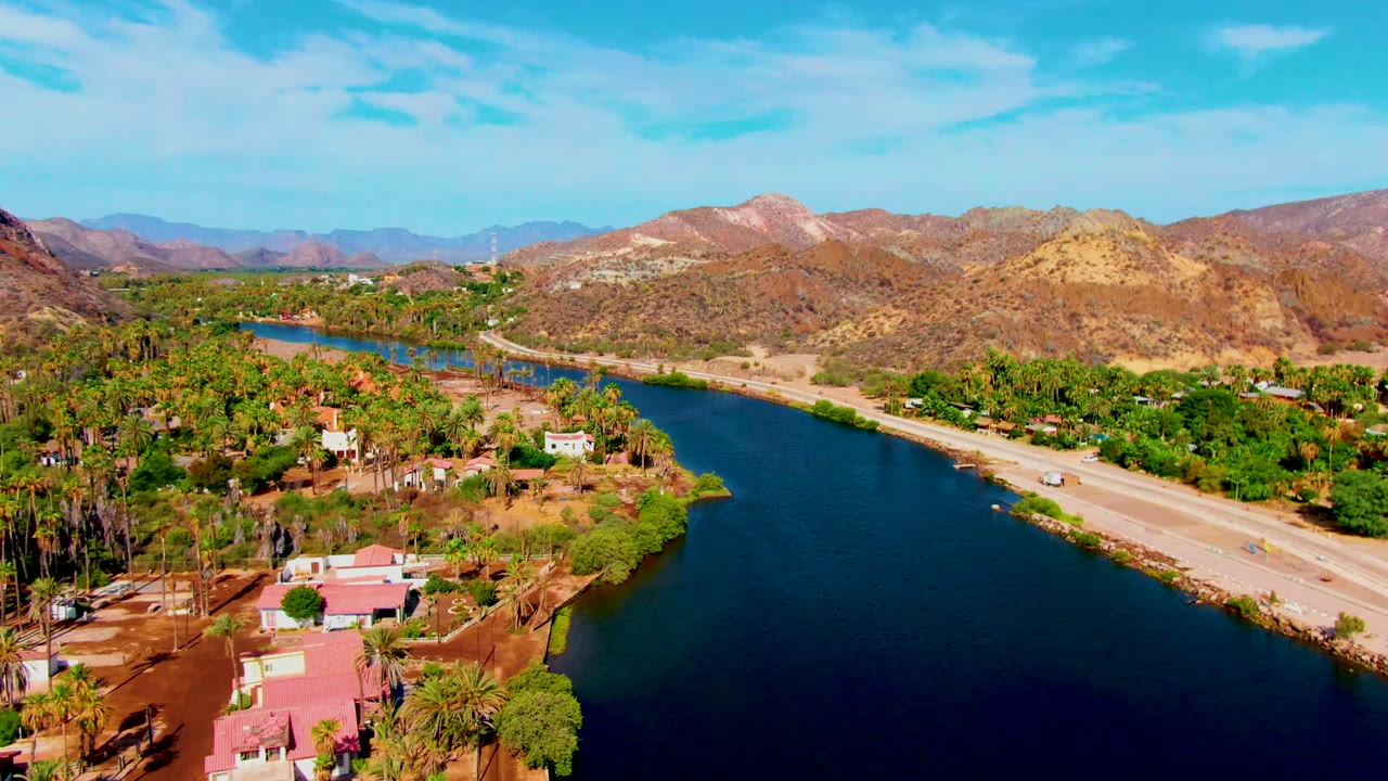 Wide aerial view of blue river water beside a road and palm-covered settlement.