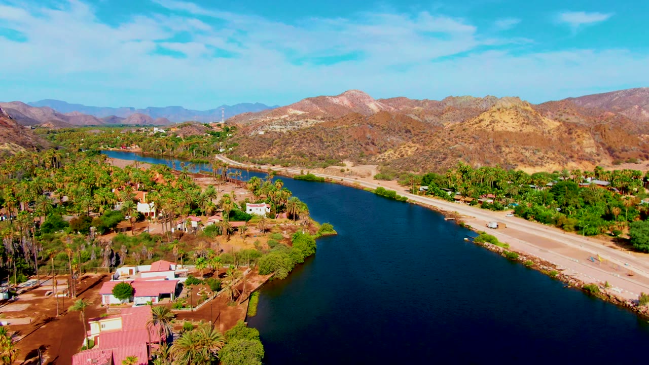 The frame shows Mulege from above with a broad blue river at right, a paved road with small vehicles, palm clusters, scattered houses, arid hills, and no visible people.