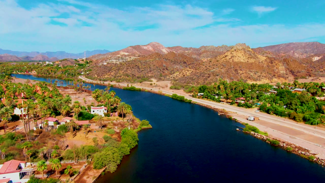 Aerial view in Mulege, Baja California Sur, with a dark canal in the foreground, a parallel road, palm trees, small houses, dry hills, and no visible people.