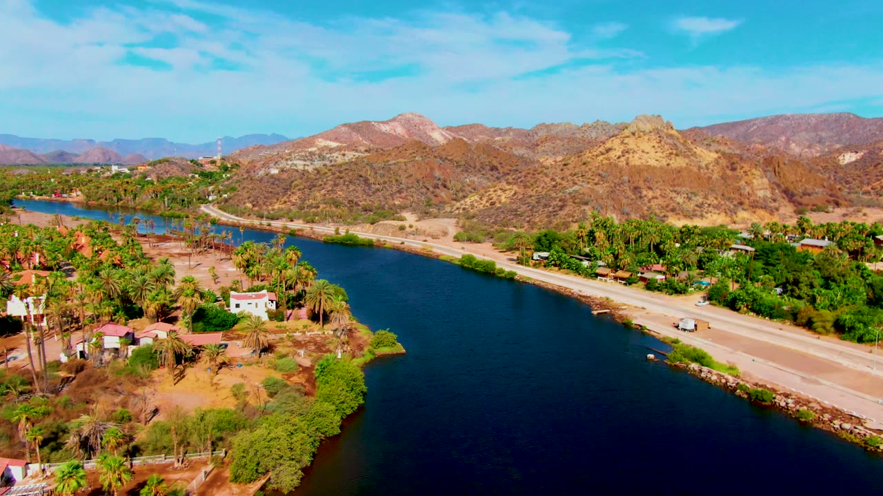 Canal and road through a palm-lined settlement with mountains behind.