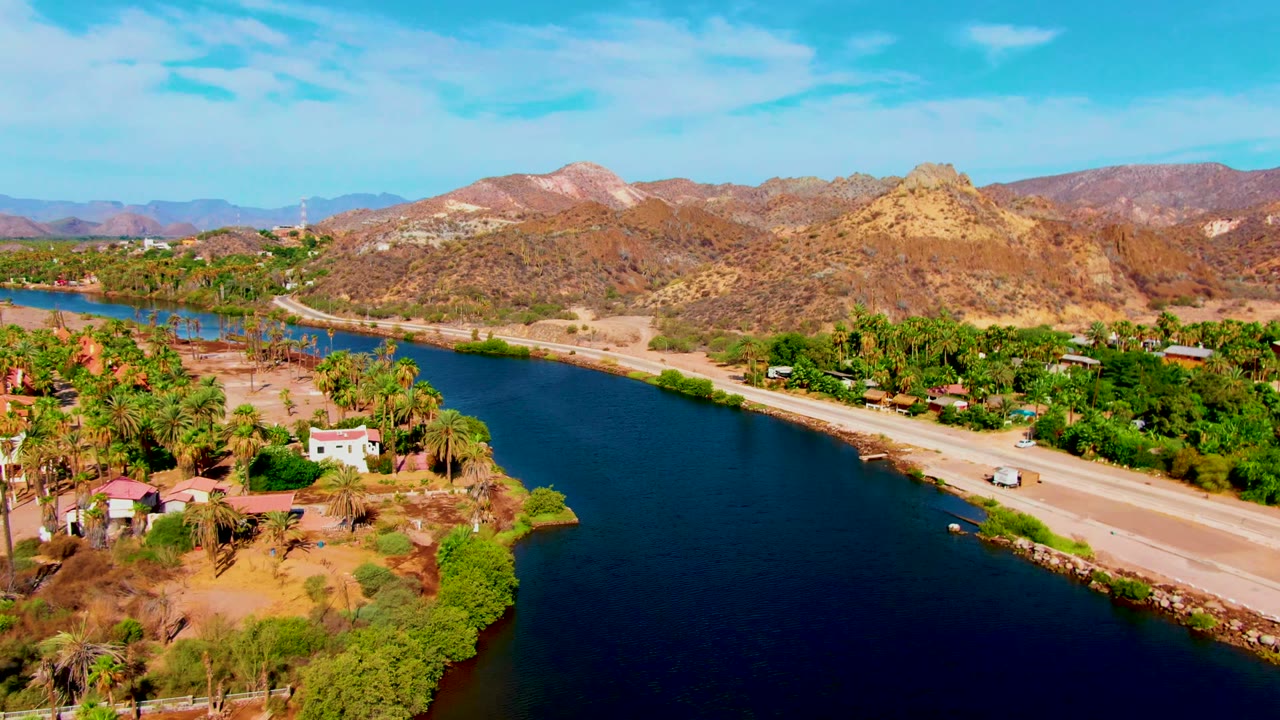 Aerial frame over Mulege, Baja California Sur, with deep blue canal water, a pale road, scattered houses, palm groves, rocky tan hills, and no visible people.