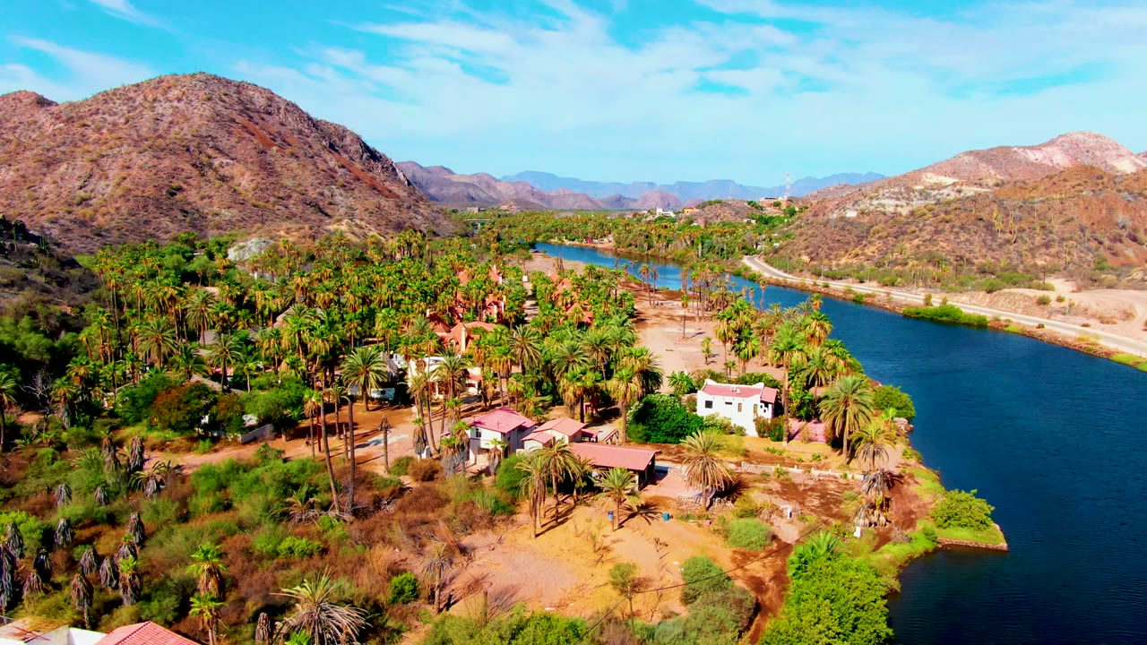 Opening aerial frame over Mulege, Baja California Sur, showing a blue river, dense palms, dry ground, small riverside buildings, rocky hills, and no visible people.