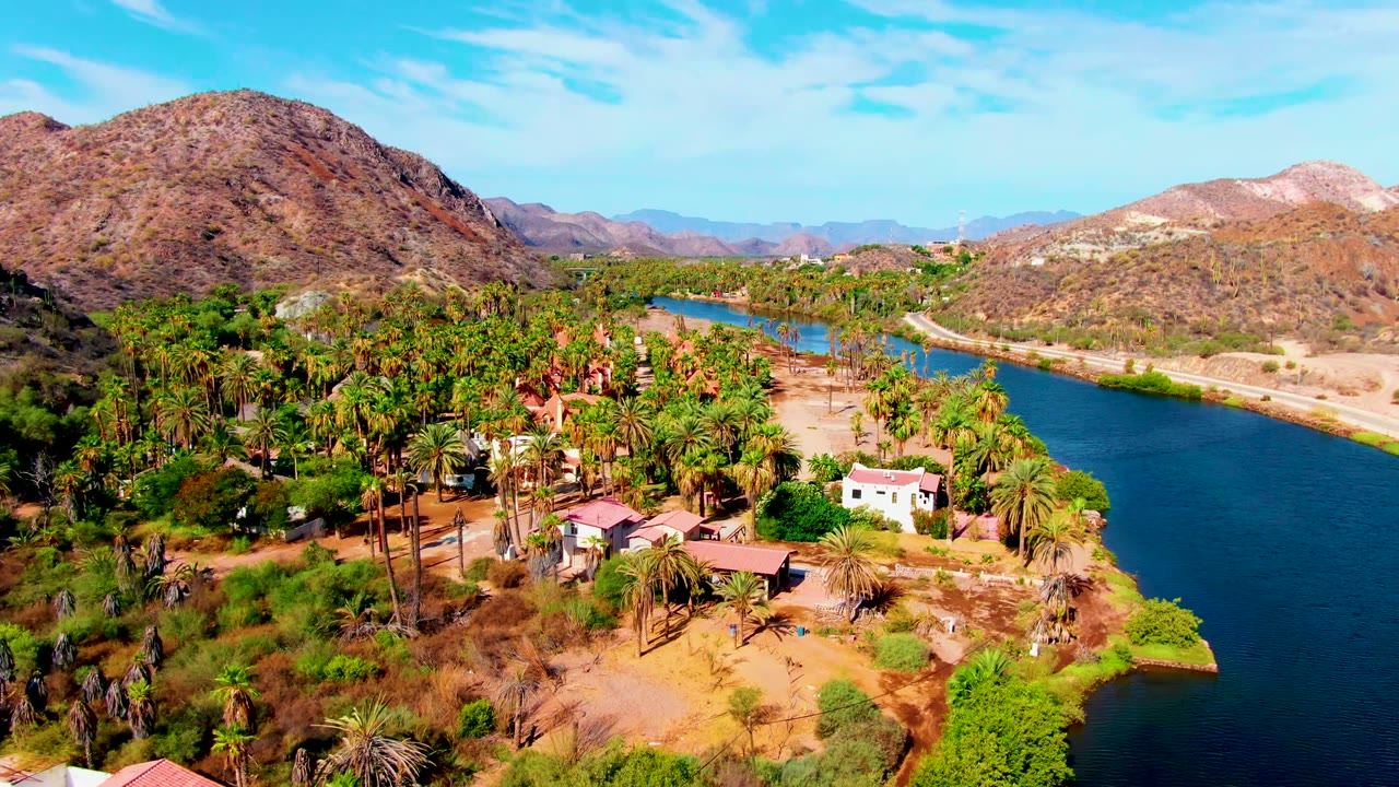 Aerial frame over Mulege showing the river on the right, palms and small structures below, rocky desert slopes across the background, and no visible people.