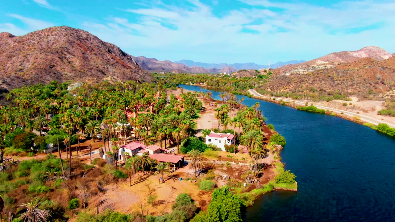 Aerial frame over Mulege, Baja California Sur showing a blue river along palm trees and small buildings, with dry hills in the background and no visible people.