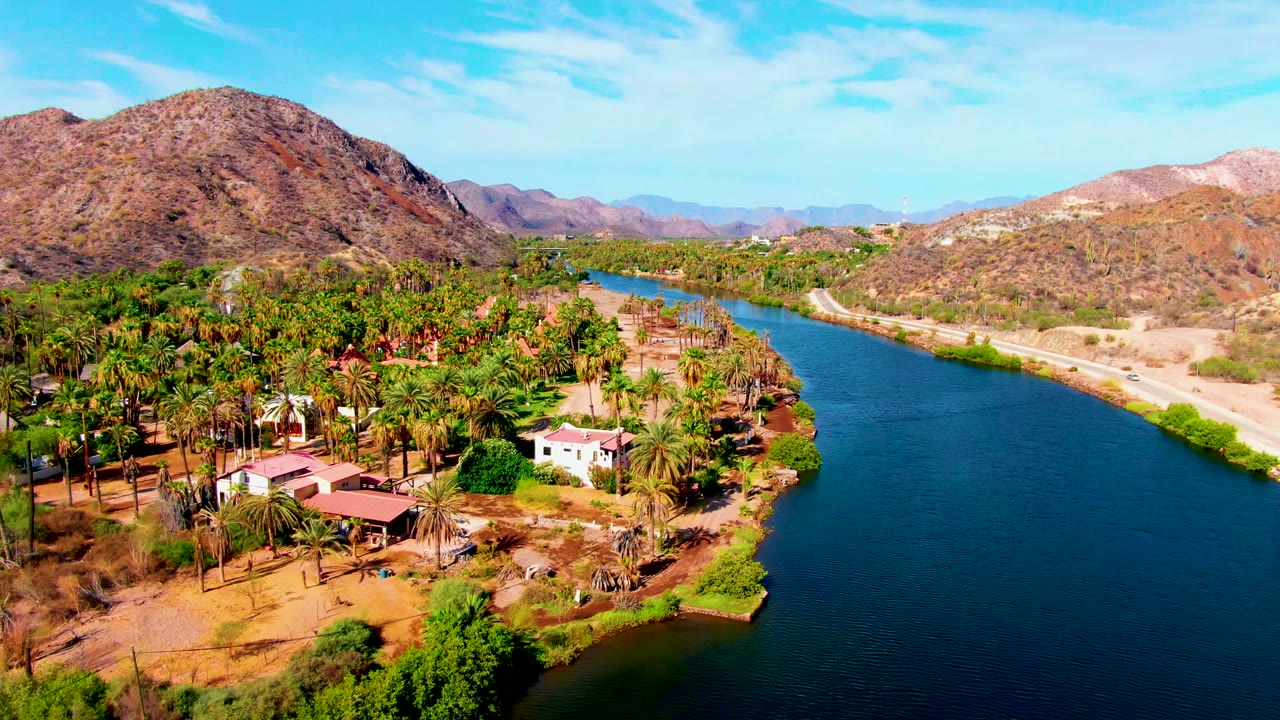 Opening aerial frame over Mulege, Baja California Sur, with a blue river curving past palm groves, scattered houses, a pale riverbank road, dry mountains, and no visible people.
