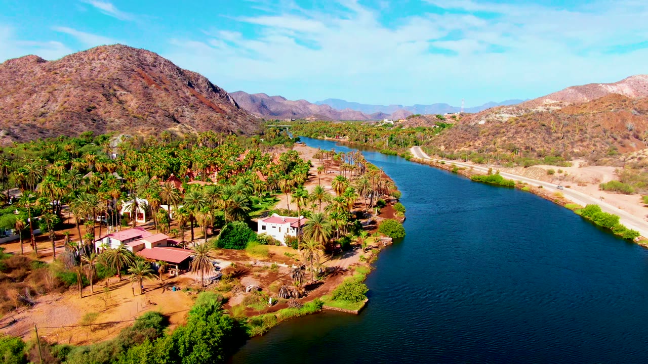 Aerial riverbank view with palms and houses in front of dry hills.