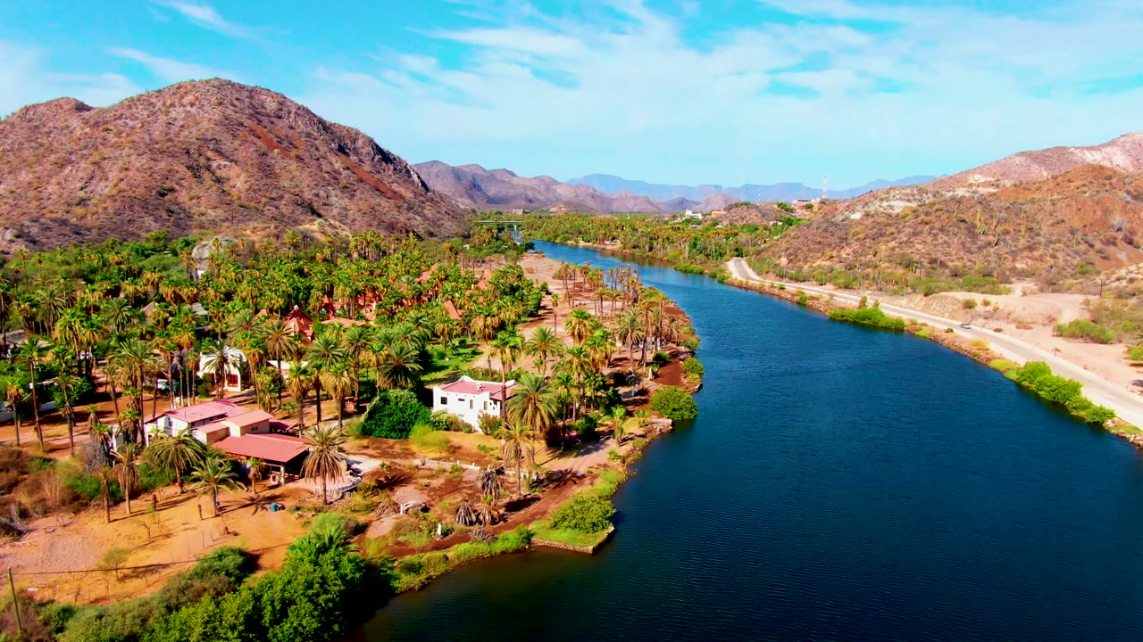 Mulege aerial frame with the blue river leading toward the distance, palms and houses along one bank, a light road on the other bank, brown mountain slopes, and no visible people.