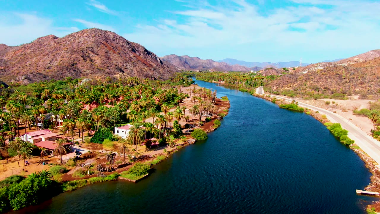 Aerial view in Mulege, Baja California Sur showing a blue river curving past palm-lined homes, a pale road, dry mountains, and no visible people.