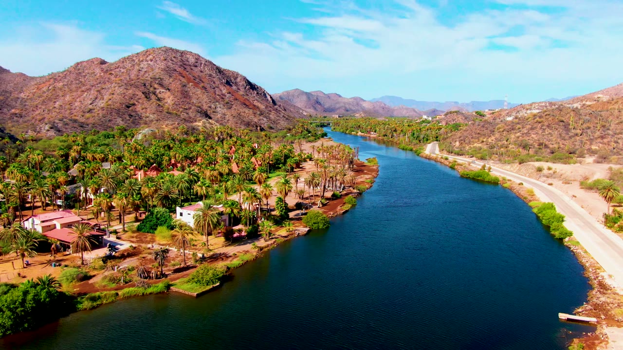Wide aerial view over Mulege with dark blue water in the foreground, green palms and homes along the bank, a light road, distant hills, and no visible people.