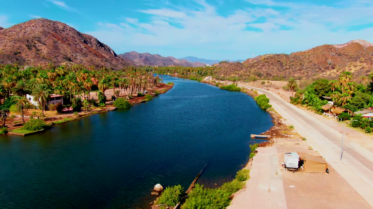 Aerial view in Mulege, Baja California Sur, Mexico, showing a blue river curving beside a pale road, palms on the left bank, dry mountains beyond, and no visible people.