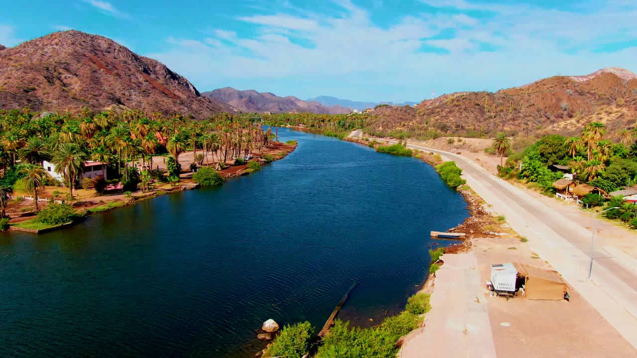 Aerial view over Mulege with textured blue water, a light-colored road on the right bank, palm trees and buildings across the river, dry mountain slopes, and no visible people.