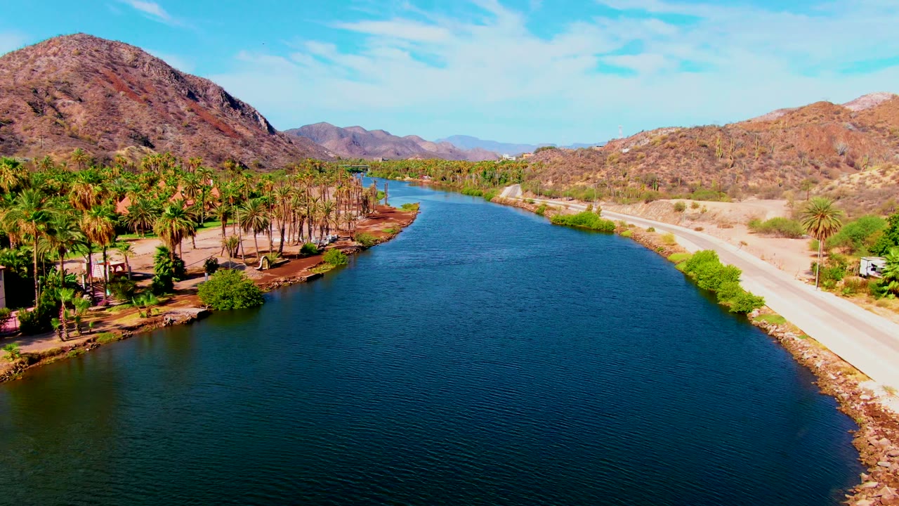 Opening aerial frame over a Mulege canal with blue water in the foreground, palm trees on the left bank, a pale road on the right bank, dry mountains behind, and no visible people.