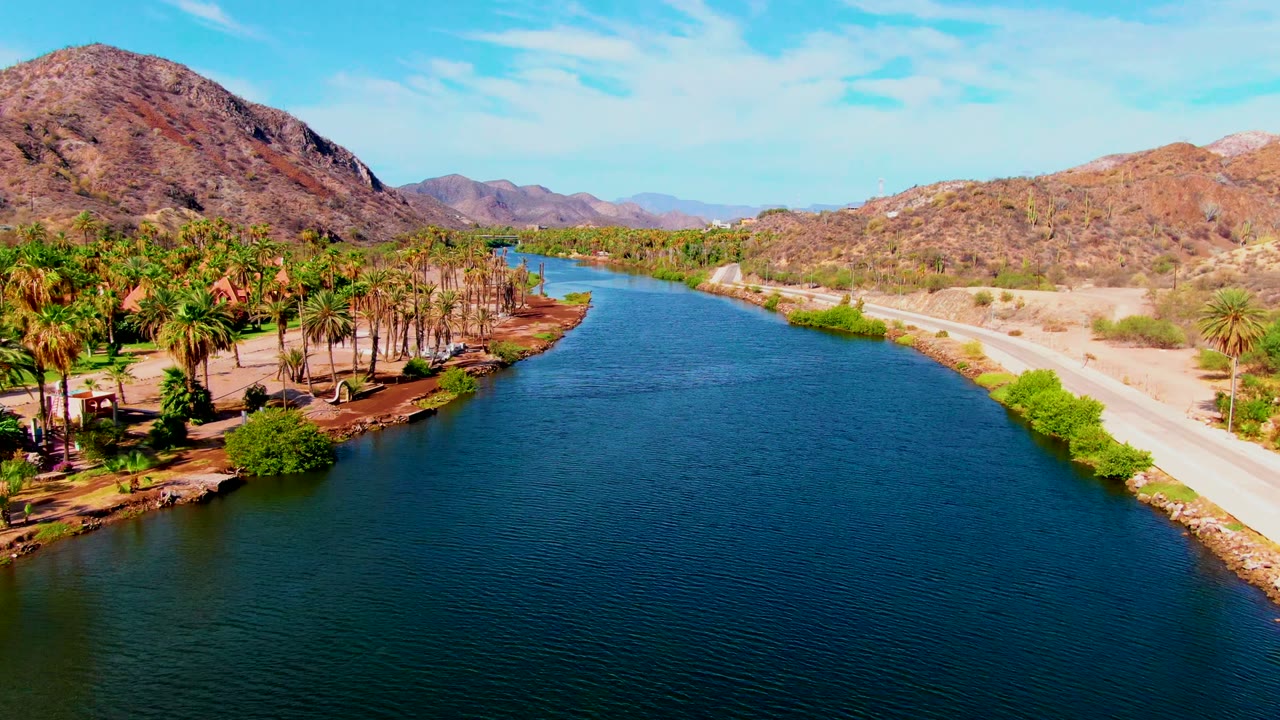 Second frame of the Mulege canal showing rippled blue water, denser palms along the near bank, the road tracing the opposite side, arid hills in back, and no visible people.