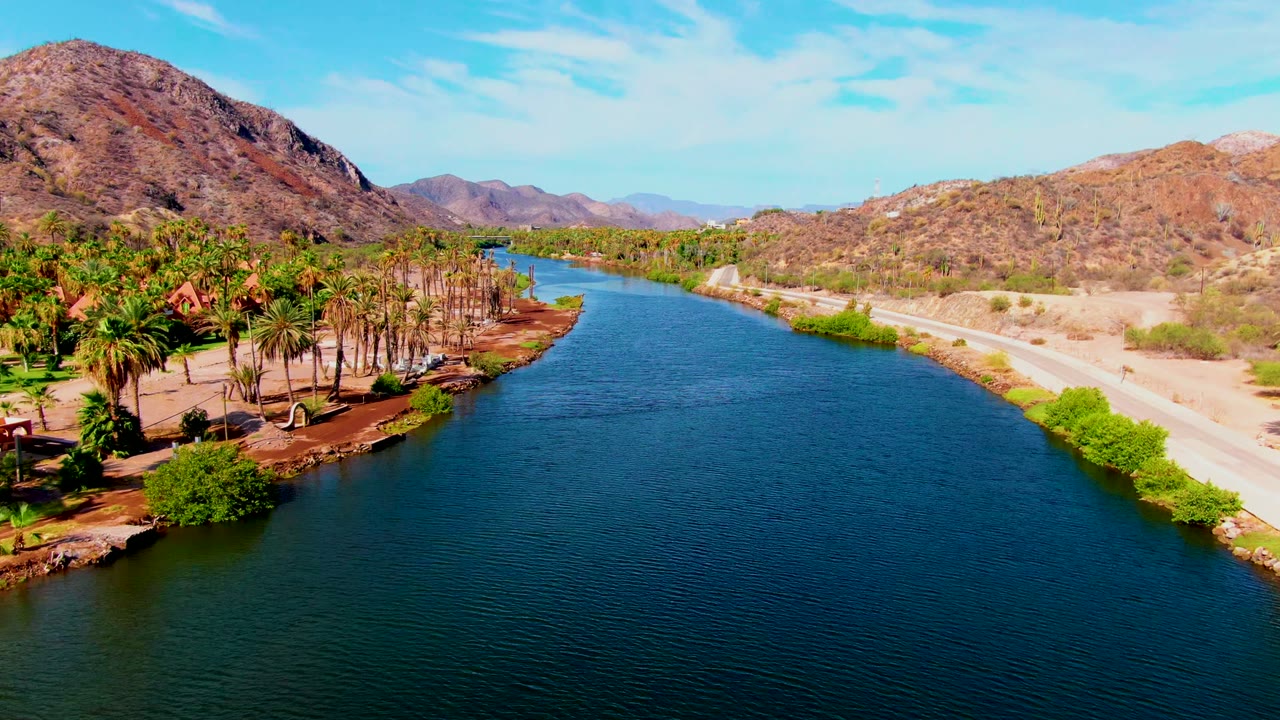 Third frame over Mulege with the canal widening across the foreground, a palm cluster on the left bank, a road and scrubby desert on the right, mountains beyond, and no visible people.