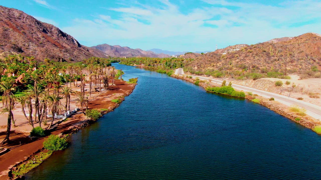 Opening frame of Mulege, Baja California Sur showing a blue river between a palm-lined sandy bank and a road below dry mountains, with no visible people.