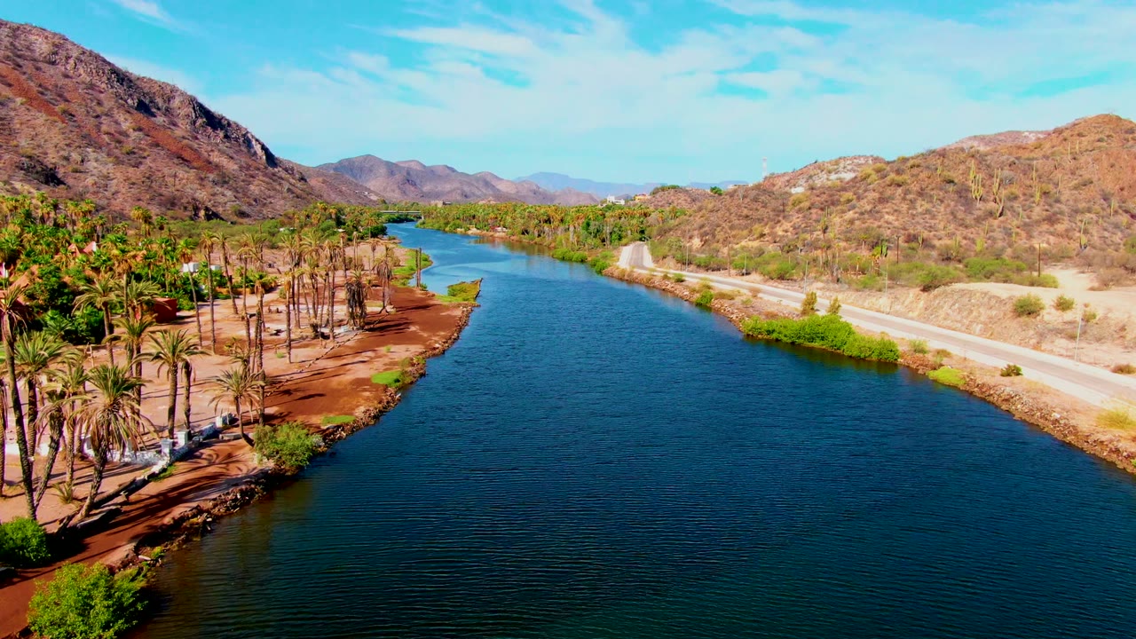 Blue river curving past palm trees and a road in dry hills.