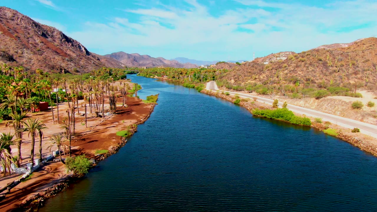 Mulege aerial view showing rippled blue water, a palm grove on reddish sandy ground, a road and embankment on the opposite bank, dry mountains, and no visible people.