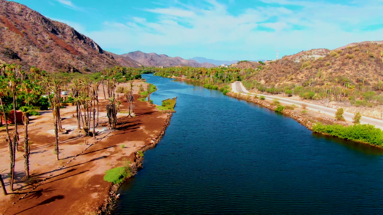 Opening aerial frame over a blue river in Mulege, Baja California Sur, with palm trees on the left bank, a pale road on the right bank, brown mountains behind, and no visible people.