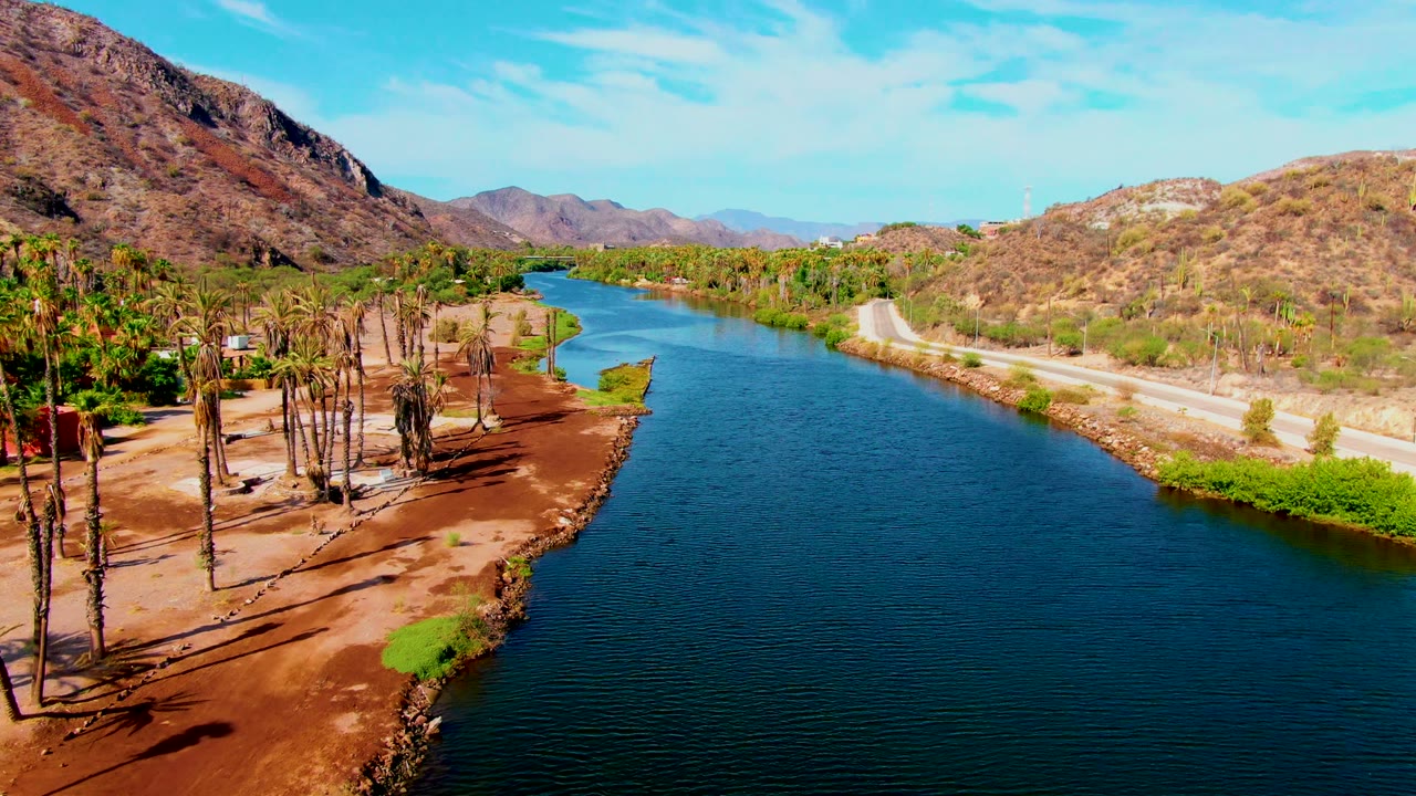 Wide aerial river bend with palms on the near bank and road along dry hills.