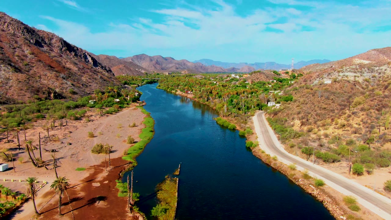 Aerial view in Mulege, Baja California Sur, showing a dark river curving past a dirt road, sparse desert vegetation, palms near the bank, rocky hills, distant mountains, and no visible people.