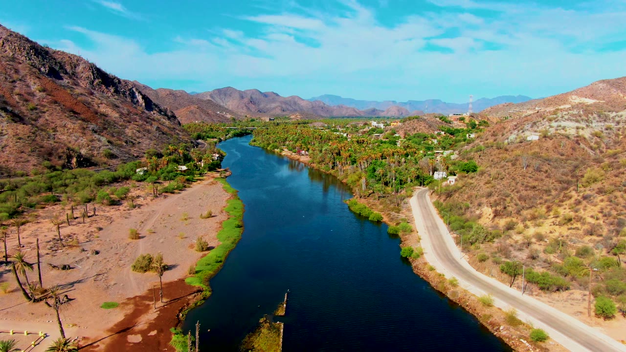 Wide aerial of blue river water, palms, road, and rocky desert slopes.