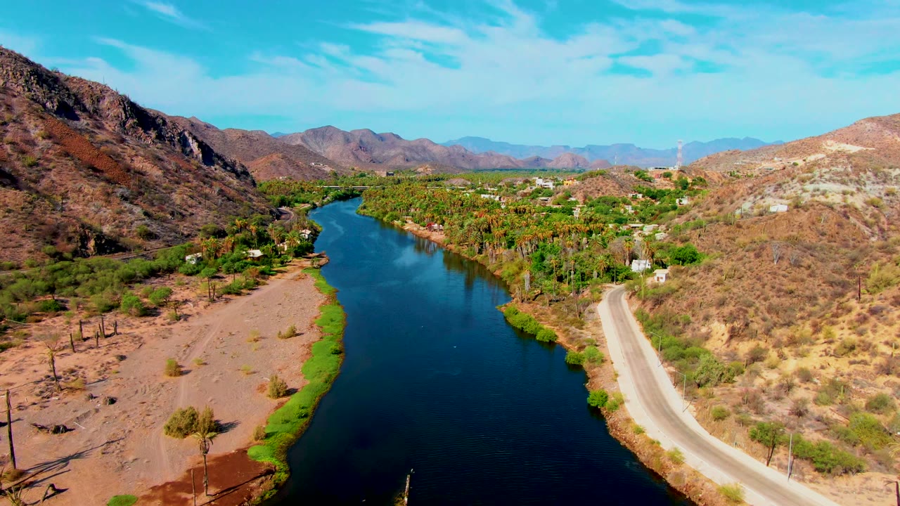 Drone-height view in Mulege, Baja California Sur, showing the river bend deeper in frame, the road tracing the right side, dry hills around the valley, palms along the water, and no visible people.