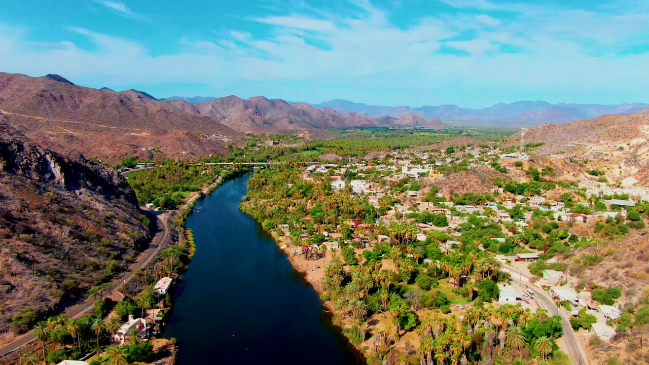 Opening aerial frame over Mulege, Baja California Sur, with the river on the left, green vegetation and buildings in the valley, brown mountains beyond, and no visible people.