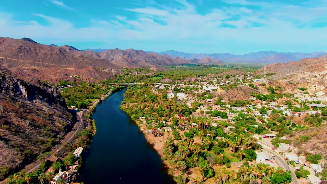 Wide aerial over river, greenery, roads, buildings, and desert hills.