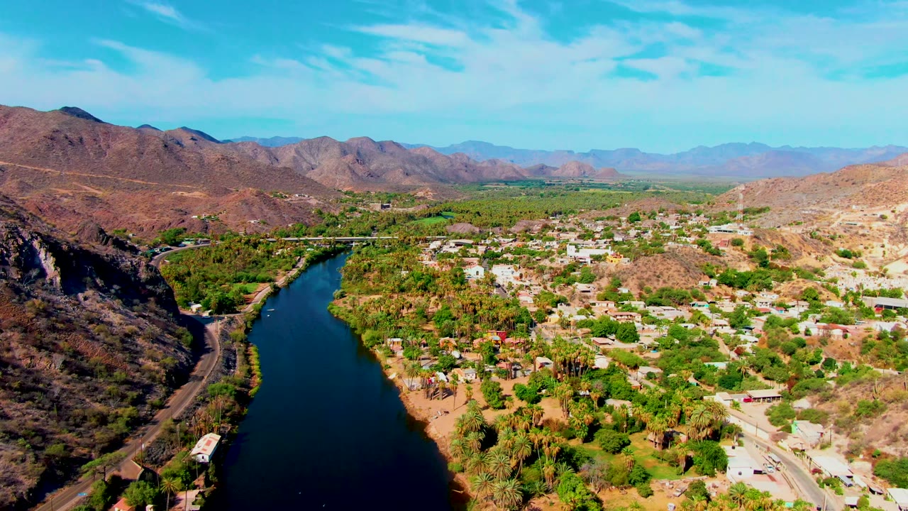 The frame looks across Mulege from above, with the river running near the left edge, roads and small vehicles below, clusters of buildings among greenery, desert ridges, and no visible people.