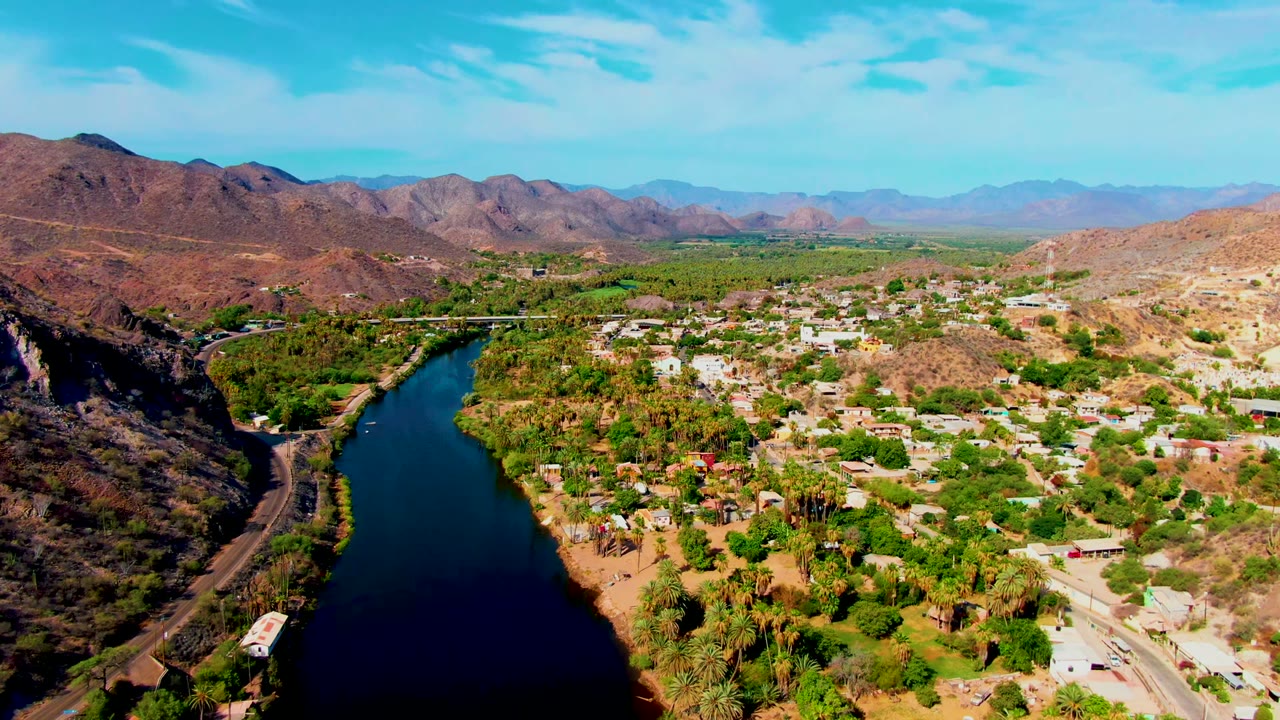 Aerial view over Mulege, Baja California Sur, with a dark river at left, green trees, pale buildings, dry hills, distant mountains, and no visible people.