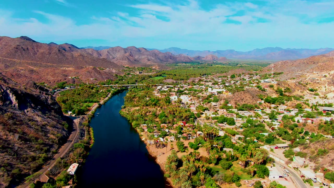 Aerial view of Mulege with the dark river curving below, dense green vegetation near the water, scattered homes and roads, dry hills, hazy mountains, and no visible people.