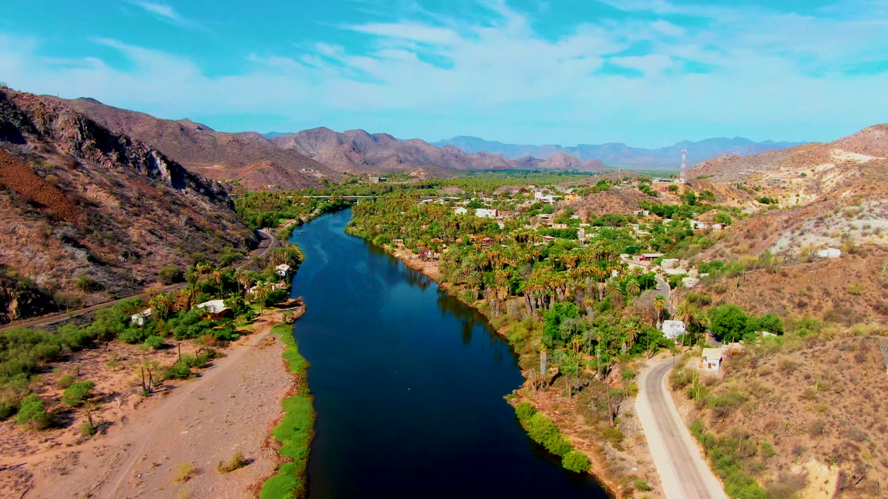 Opening aerial frame over Mulege, Baja California Sur, showing a dark river curving between dry hills, green banks, scattered houses, a road, distant mountains, and no visible people.