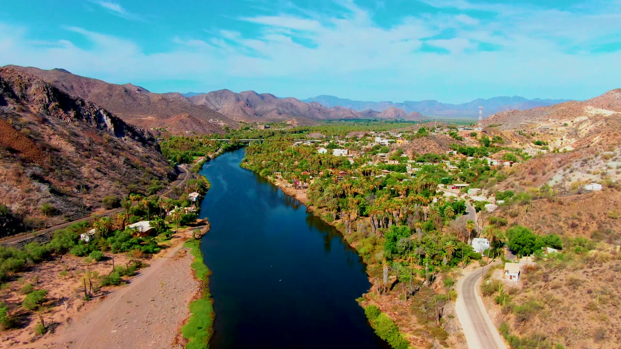 Aerial frame in Mulege with the river centered left, a pale road on the near bank, palms and small buildings in the valley, dry hills behind, and no visible people.